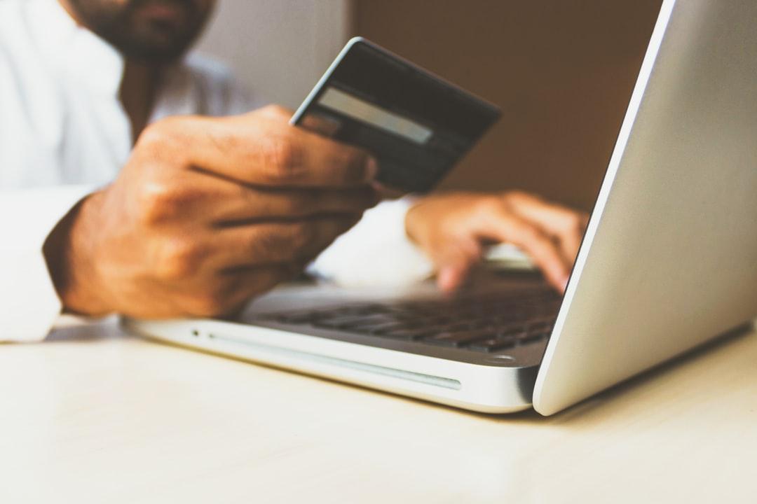 A person typing on a laptop with one hand, and holding a credit card in the other.