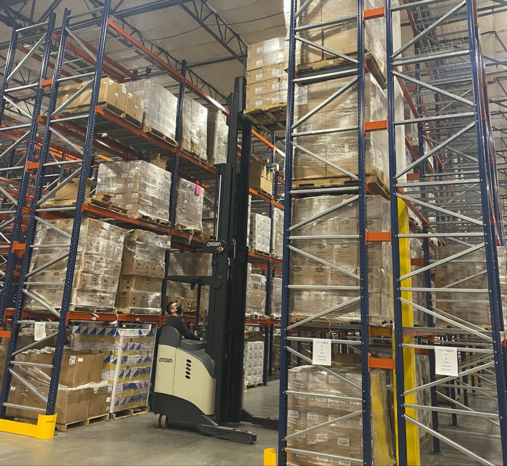 A person operating a lift to take cardboard boxes off of shelves in a warehouse.