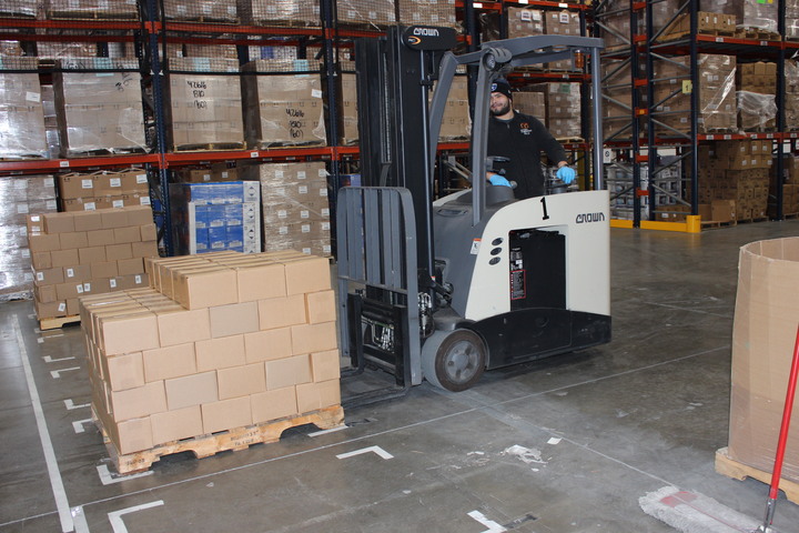A person operating a small forklift in a warehouse, lifting several boxes.
