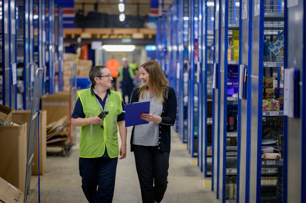 Two people walking and chatting together in a warehouse, one holding a clipboard.