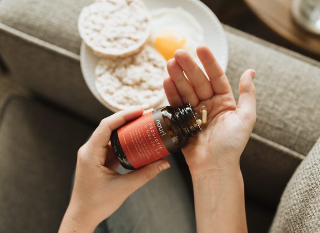 A person pouring 2 pills out of a supplement bottle into their palm, with a plate of rice cakes and eggs below them.