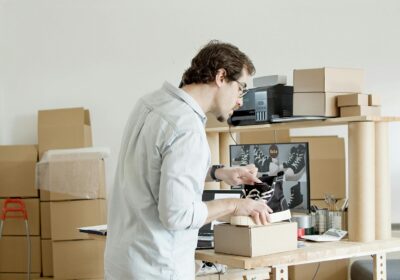 A man surrounded by cardboard boxes, packing up a pair of black and white, casual sneakers.