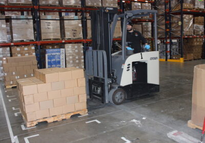 A person operating a small forklift, lifting multiple cardboard boxes in a warehouse.