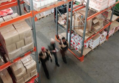 A group of three people walking together in a 3PL warehouse, surrounded by shelves of cardboard boxes.